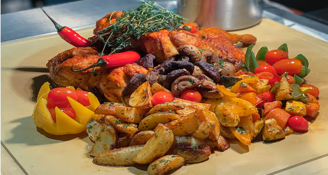 Roasted chicken garnished with thyme and red chili peppers, served with seasoned potato wedges, grilled mushrooms, cherry tomatoes, and mixed bell peppers on a beige platter.