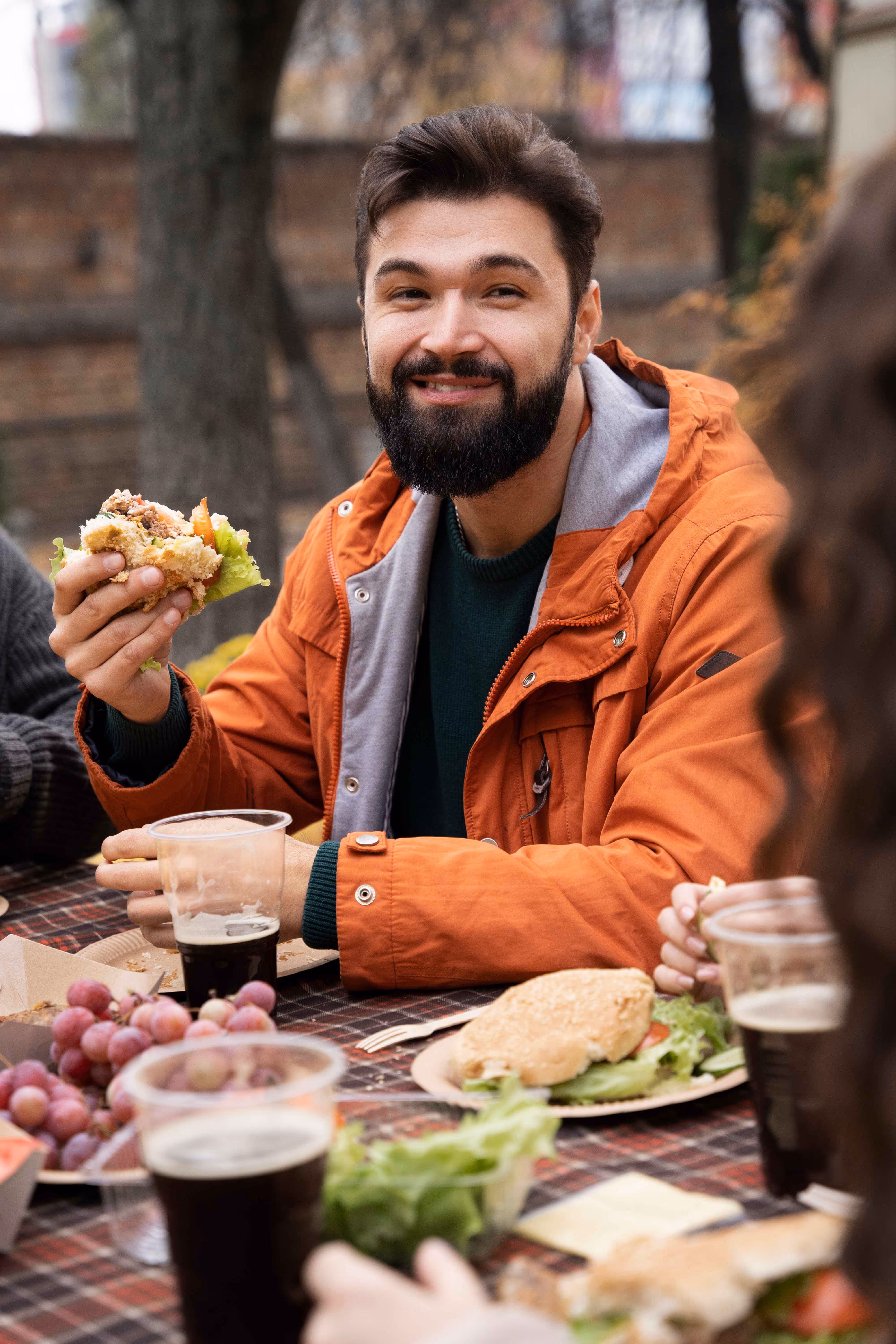 Smiling man in an orange jacket eating a sandwich outdoors at a picnic table with friends.